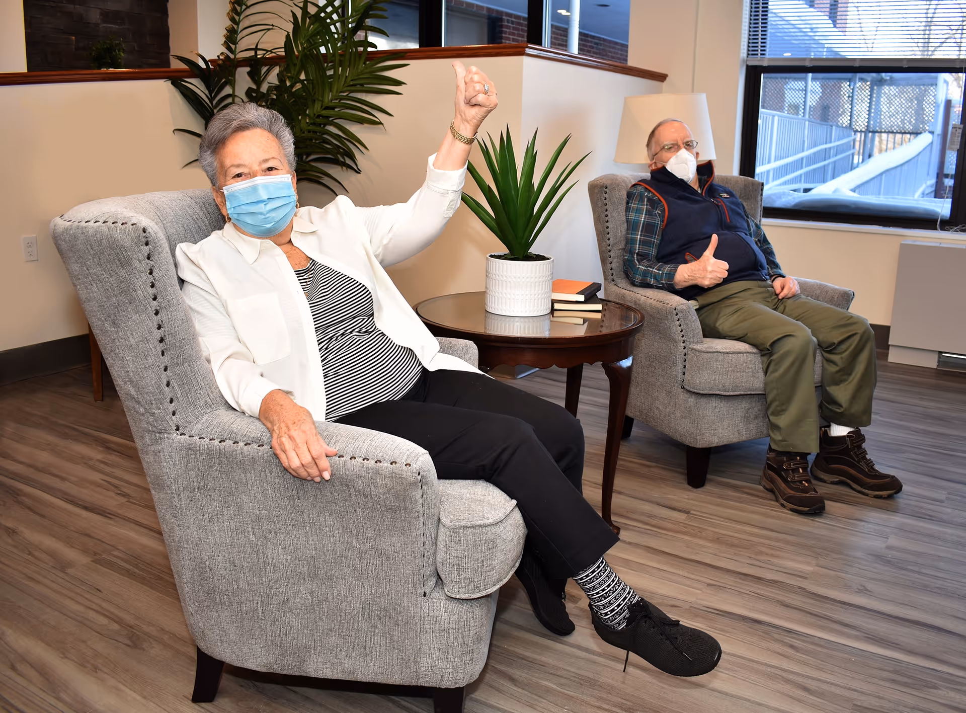 Two elderly individuals wearing face masks sitting in gray upholstered armchairs in a well-lit room with wooden flooring. They are giving thumbs-up gestures. A round wooden table with a potted plant and some books is placed between them. There is a large window in the background letting in natural light.