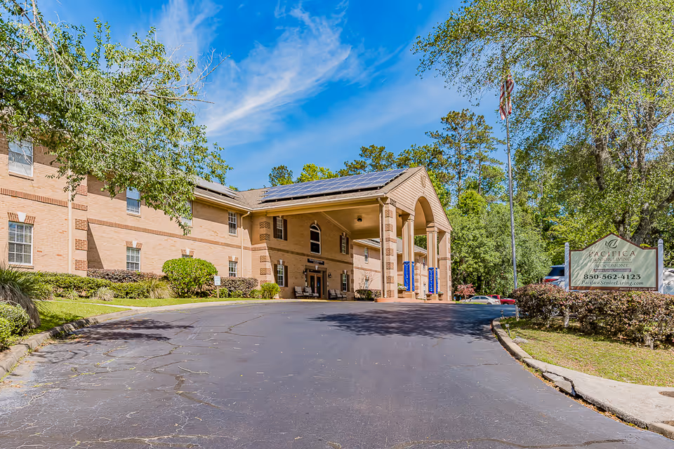 Exterior view of Woodmont Senior Living facility showing a two-story brick building with a covered entrance supported by columns. The driveway leads up to the entrance, and there are trees and bushes surrounding the area under a blue sky with some clouds. A sign near the entrance reads 'Pacifica Senior Living Woodmont' with a phone number and website.
