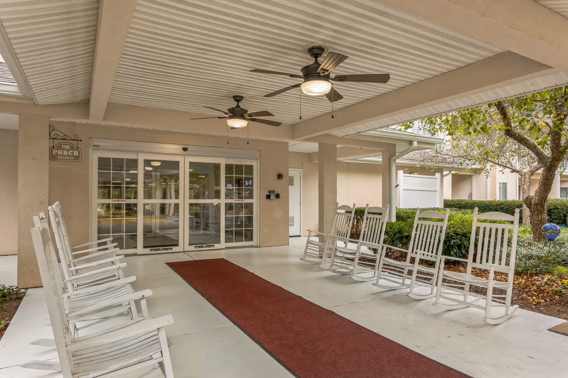 Covered entrance porch with white rocking chairs, ceiling fans, and a red runner leading to glass sliding doors.