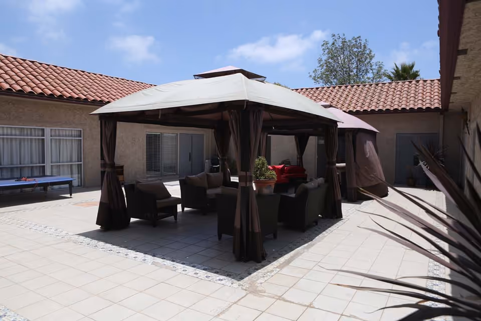 Sunlit tiled courtyard with a central covered gazebo, outdoor seating and surrounding stucco buildings with red tile roofs.