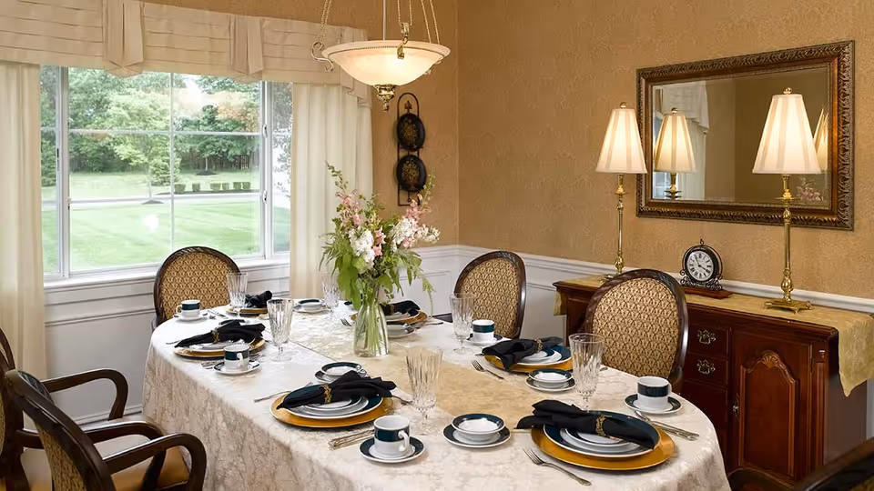 A formal dining room with an oval table set for six people. The table is covered with a white and beige tablecloth and set with plates, cups, saucers, crystal glasses, and black napkins. A vase with flowers is in the center of the table. The room has beige walls, a large window with cream curtains, a wooden sideboard with two lamps and a clock, and a large mirror above the sideboard.