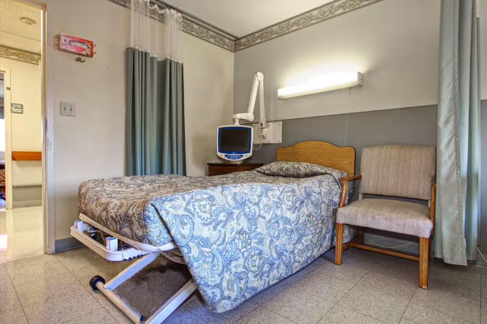 A small healthcare facility room with a single hospital bed covered in a patterned blue and beige bedspread. Next to the bed is a beige upholstered chair with wooden arms. On the wall above the bed is a mounted medical monitor and a fluorescent light fixture. The room has light-colored walls with a decorative border near the ceiling and a curtain partially covering a window on the right side. The door to the room is open, showing a hallway outside.