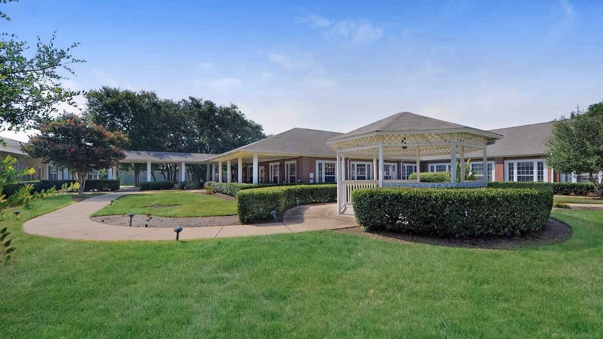 Courtyard with a white gazebo, manicured lawns, and a single-story brick senior living building with covered walkways.