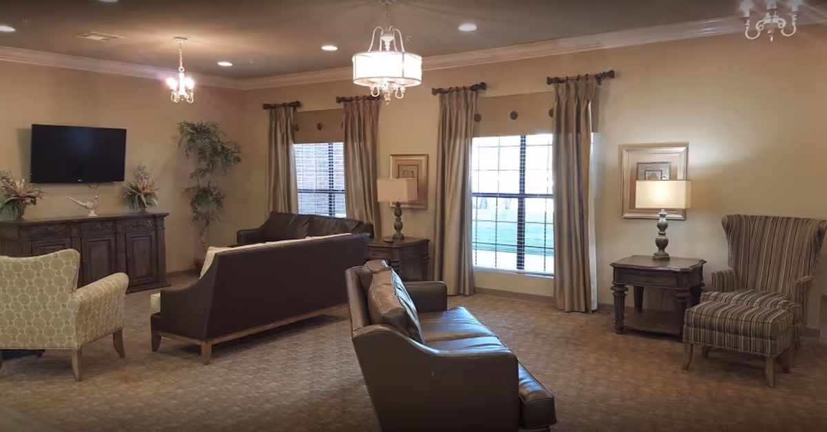 A cozy living room with beige walls and carpet, featuring two large windows with curtains, two brown leather sofas, a patterned armchair, a striped wingback chair, two wooden side tables with lamps, a wooden cabinet with decorative plants, and a wall-mounted flat screen TV.
