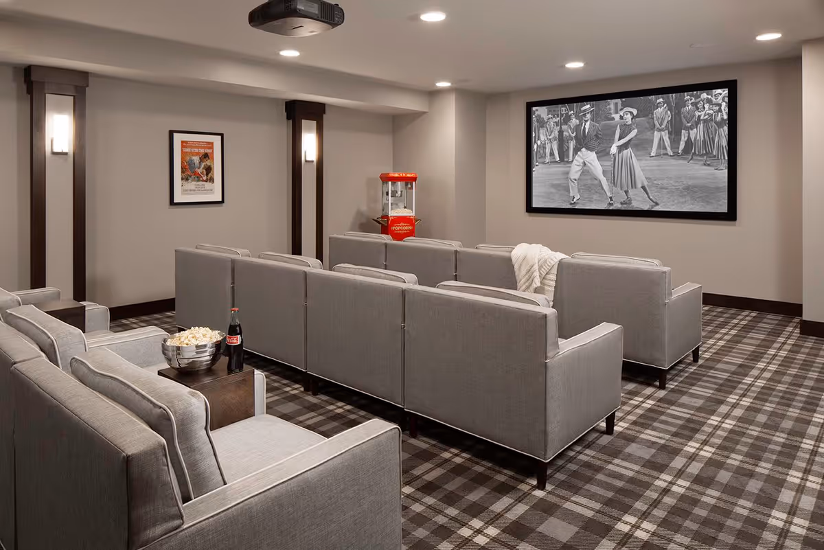 A cozy home theater room with rows of gray upholstered chairs facing a large screen showing a black and white movie scene. There is a popcorn machine in the corner, a bowl of popcorn and a bottle of soda on a small table, and a framed movie poster on the wall. The room has plaid carpet and soft lighting.