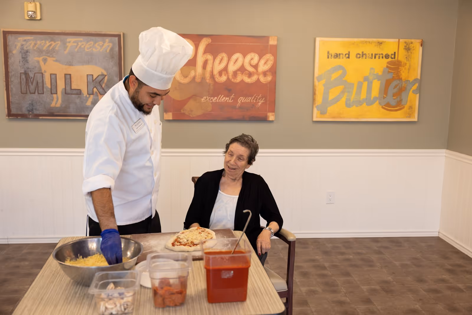 A chef wearing a white uniform and hat is preparing a pizza with cheese and tomato sauce on a table, while an elderly woman in a wheelchair watches and smiles. The background has three vintage-style signs that read 'Farm Fresh Milk', 'Cheese excellent quality', and 'hand churned Butter'.