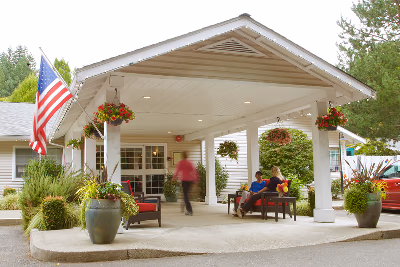 Covered entrance area of a senior living facility with hanging flower baskets and large potted plants. Two people are seated on outdoor chairs having a conversation, while another person is walking nearby. An American flag is displayed on the left side of the entrance.