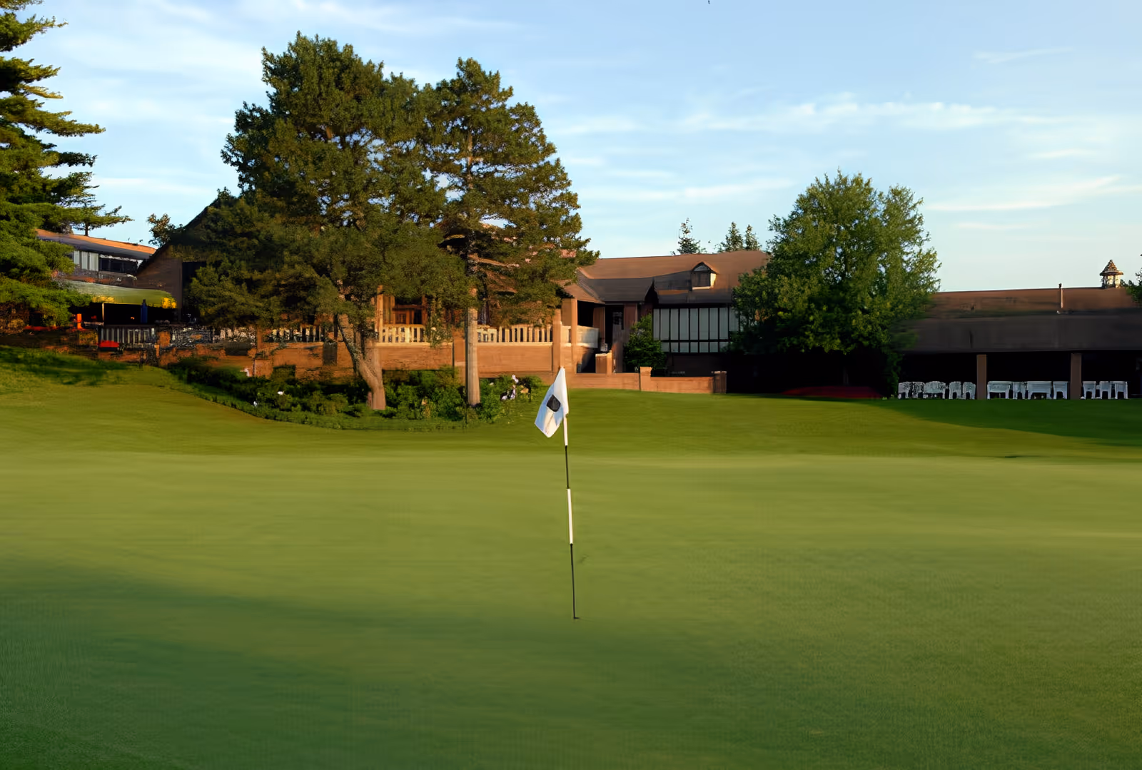 A golf green with a flagstick in the foreground and a clubhouse surrounded by trees in the background.