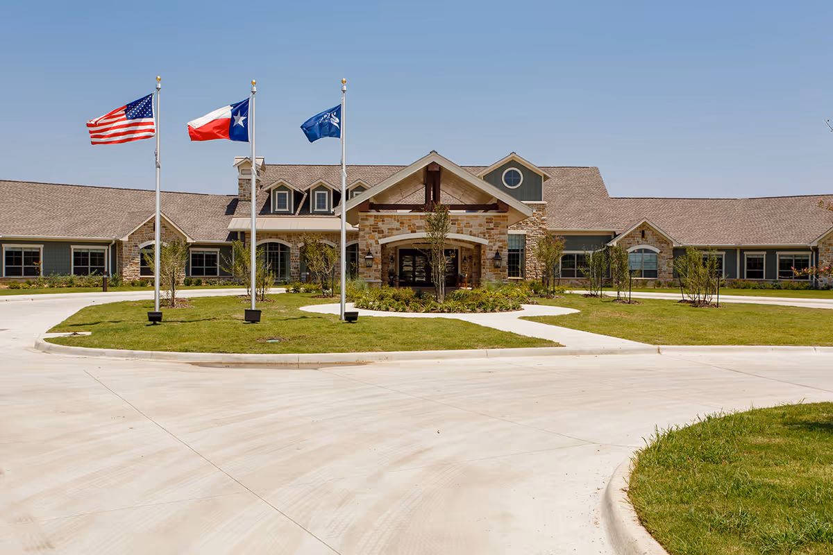 Front entrance of a single-story senior living building with three flagpoles and a circular driveway.
