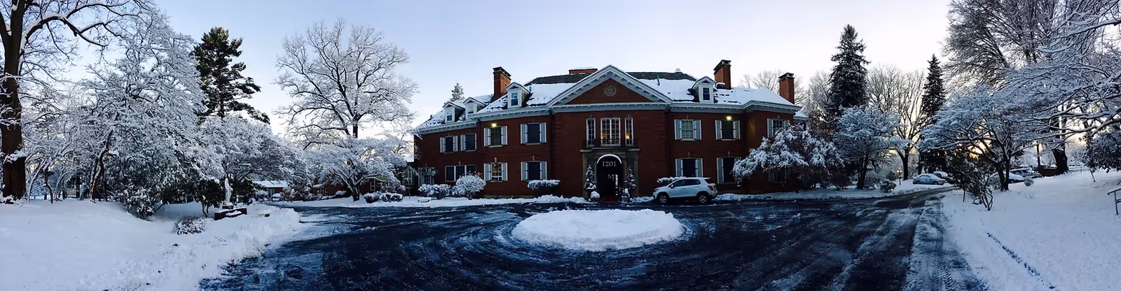 Snow-covered red-brick mansion with a circular driveway and trees in a winter landscape.