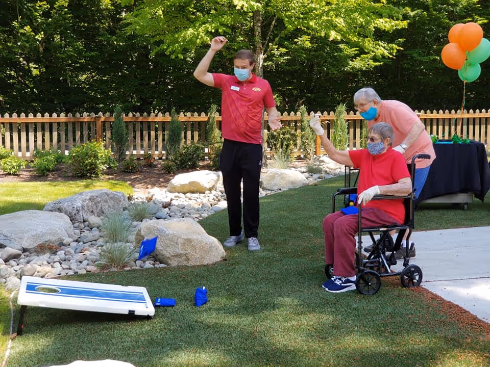 An outdoor scene at a senior living facility where two elderly individuals, one in a wheelchair and the other standing behind, are playing a game of cornhole with a younger staff member. All three are wearing face masks. The area is grassy with rocks and plants, and there is a wooden fence and trees in the background. Balloons are tied to a table nearby.