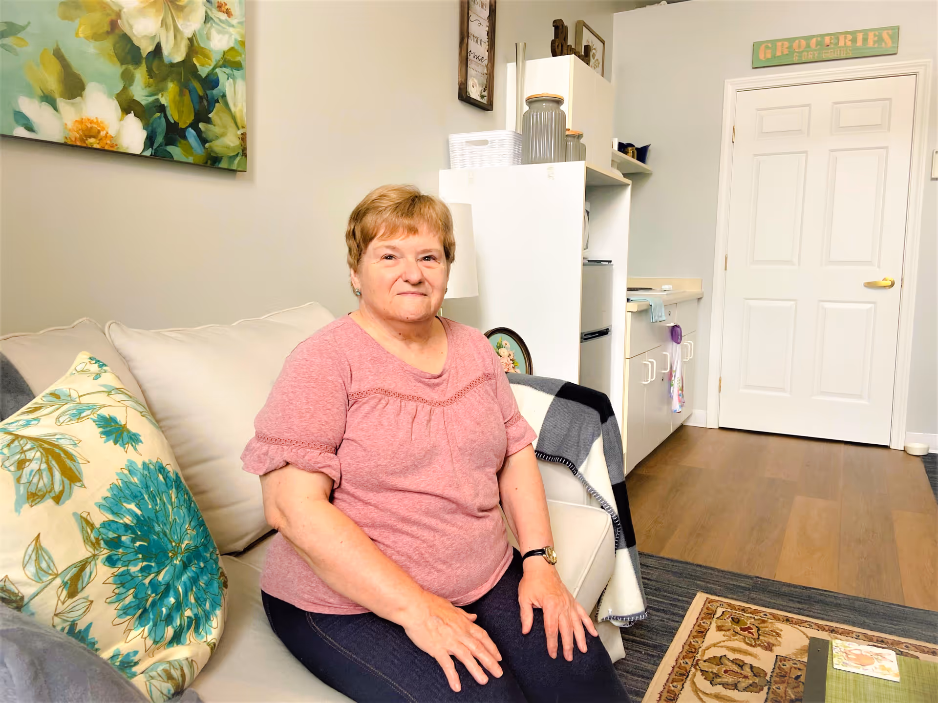 An elderly woman wearing a pink shirt and dark pants sits on a beige couch with floral and solid pillows in a cozy living space. Behind her is a white cabinet with jars and a small kitchenette area with a door labeled 'Groceries & Dry Goods'. The room has light-colored walls, wooden flooring, and a decorative floral painting on the wall.