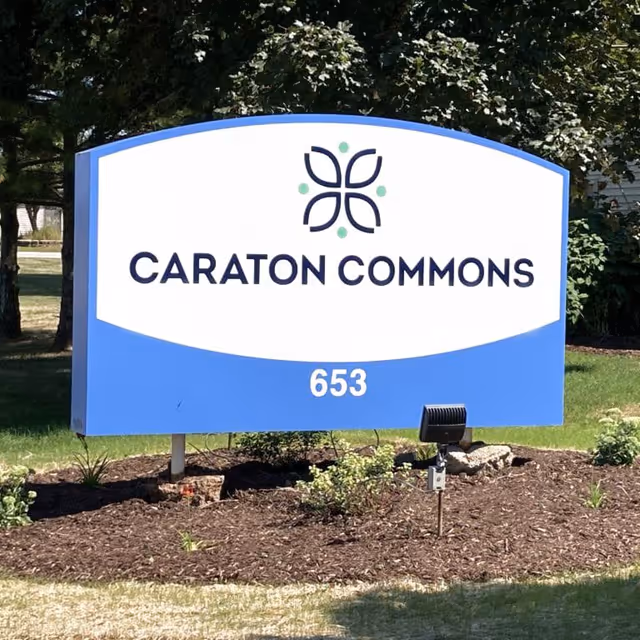 Outdoor sign for Caraton Commons with the number 653 displayed below the name, surrounded by grass, mulch, and some small plants, with trees in the background.
