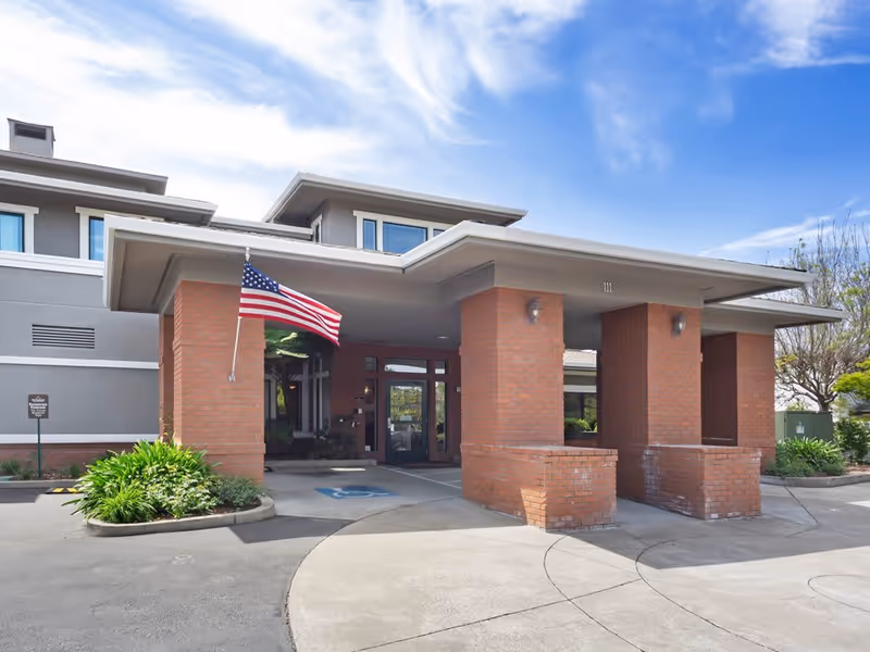 Entrance of a senior living facility with a covered drop-off area supported by brick pillars. An American flag is mounted on one of the pillars. There is a handicap parking space near the entrance, and the building has large windows and a gray exterior with white trim. The sky is partly cloudy.