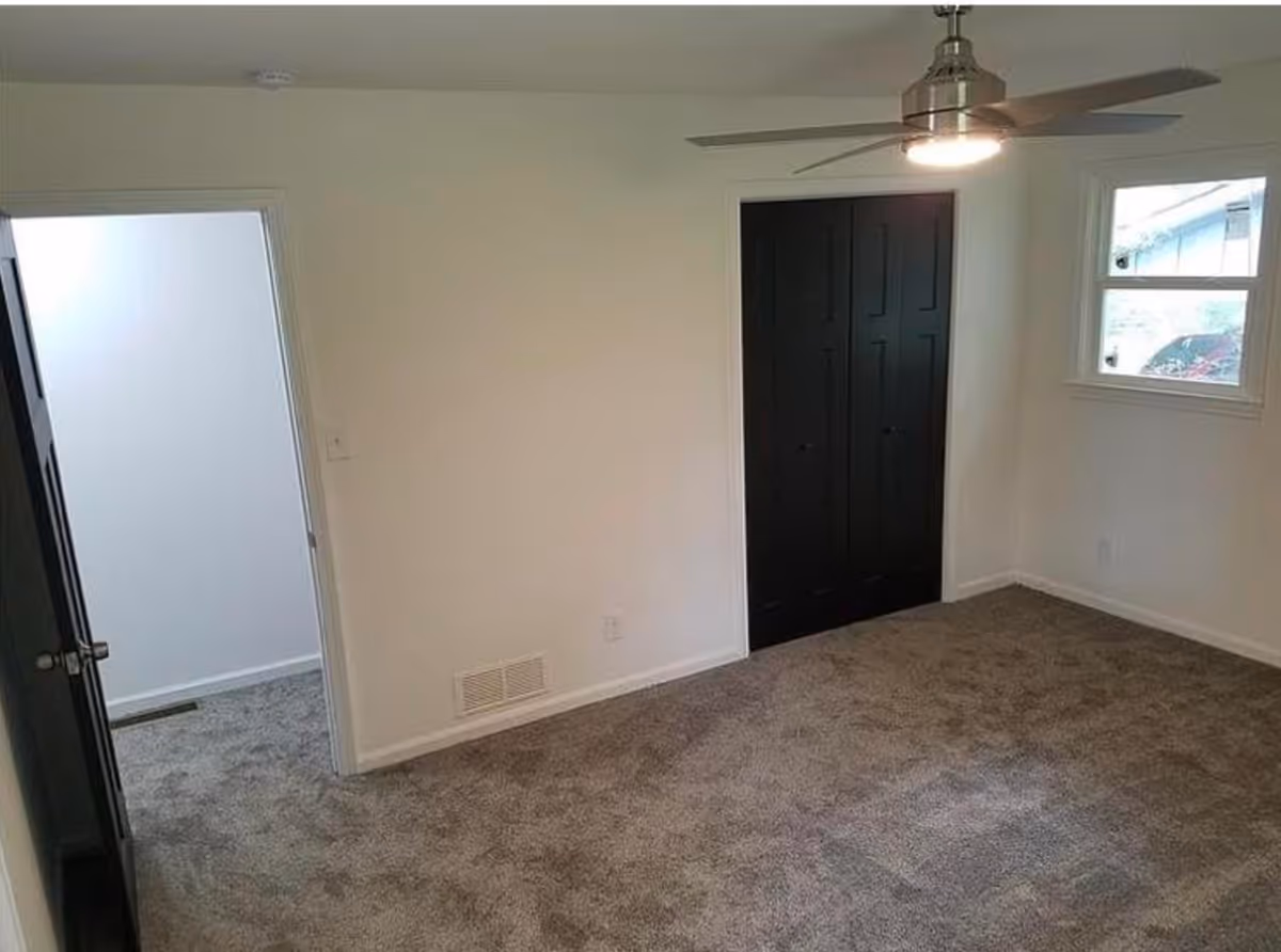 Empty carpeted bedroom with a ceiling fan, dark closet doors, and a small window.