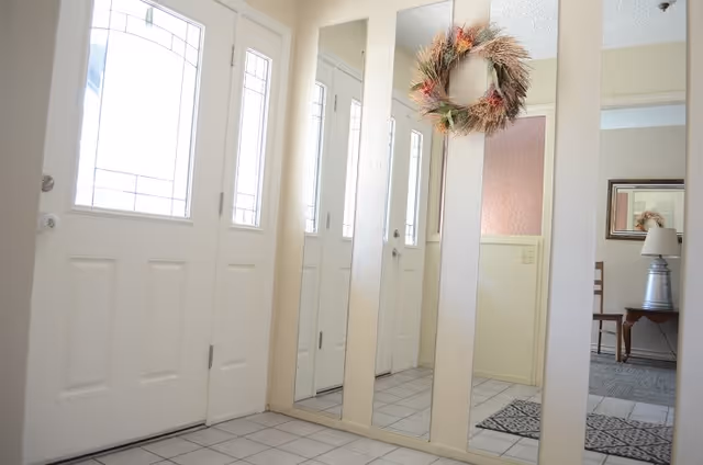 Entryway with a white front door featuring decorative glass panels, adjacent to three tall vertical mirrors on the wall. A decorative wreath hangs on one of the mirrors. The floor is tiled in white, and a glimpse of a room with a chair, table, lamp, and framed mirror is visible in the background.
