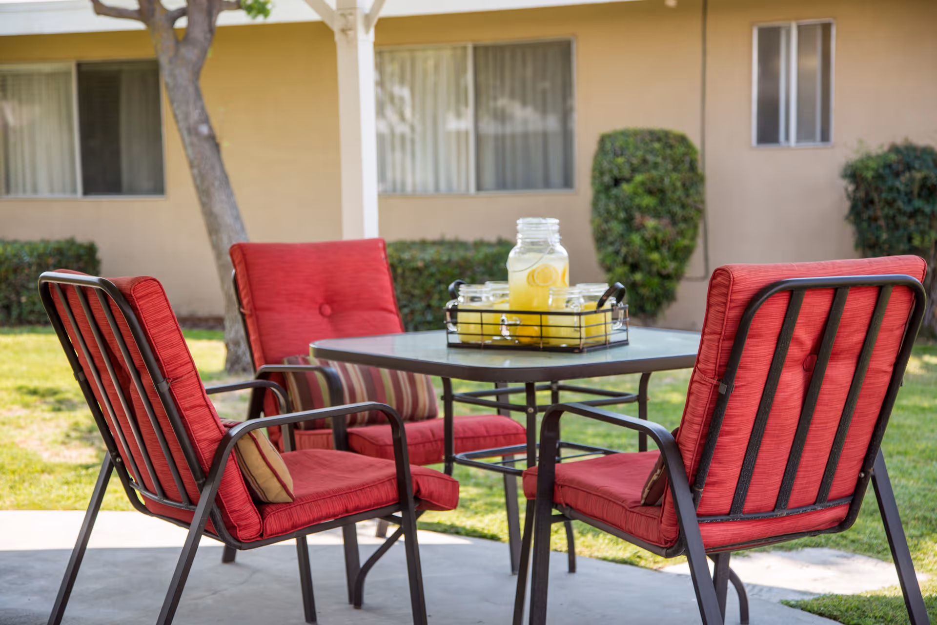 Outdoor patio area with a square table and four red cushioned chairs. On the table is a tray holding a large glass jar of lemonade and four small glasses. The background shows a beige building with windows and some green bushes.