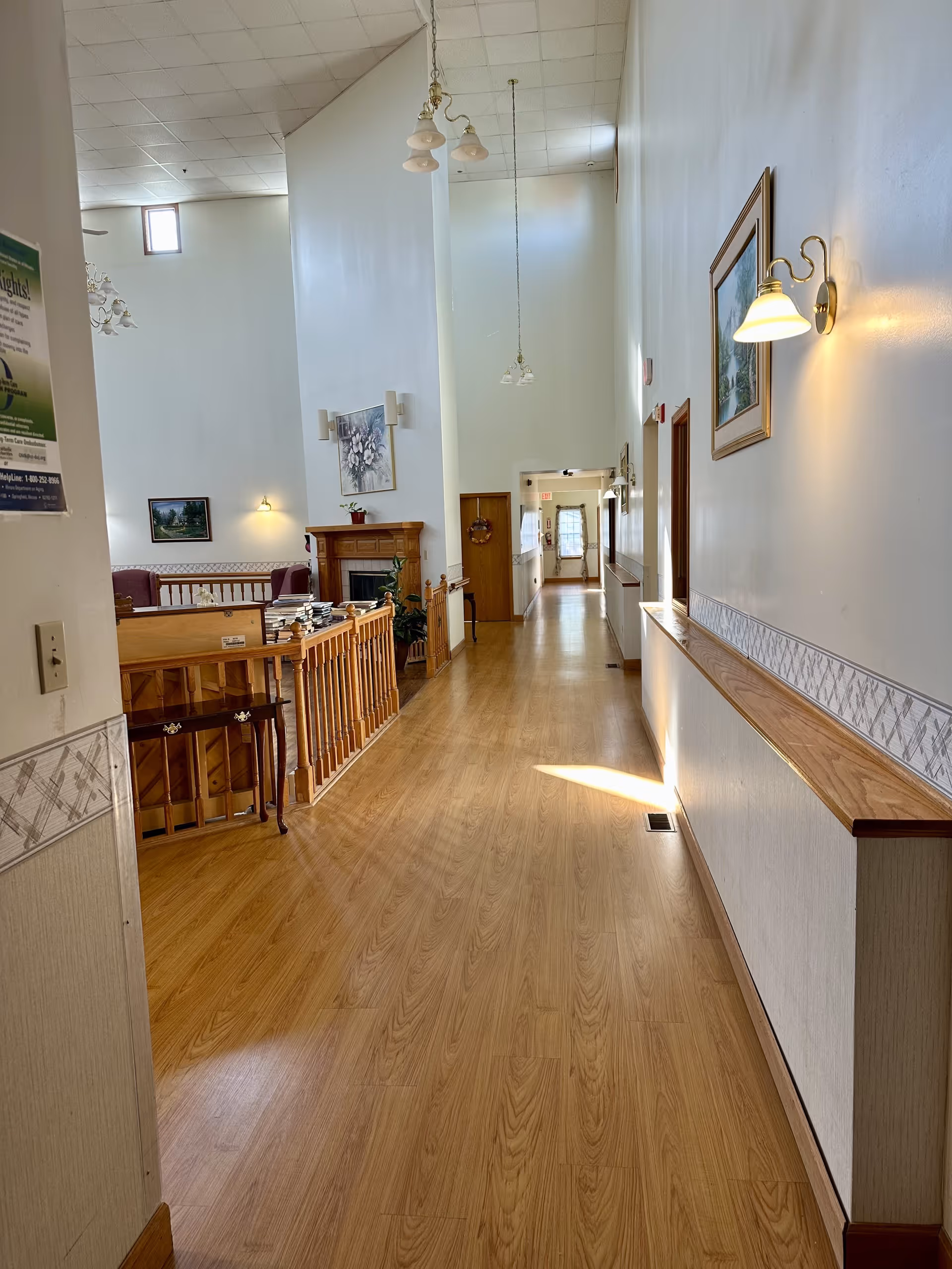 A long hallway with wooden flooring and white walls featuring framed artwork and wall-mounted lights. To the left, there is a wooden railing overlooking a lower area with a fireplace and seating. The hallway is well-lit with natural light coming from windows at the far end.
