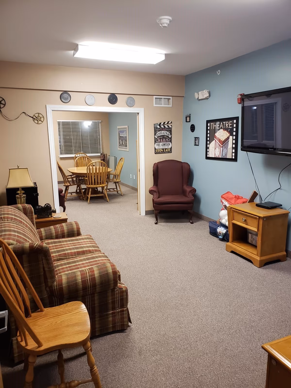 A cozy living room area with a plaid sofa, wooden chair, and a maroon armchair. A flat-screen TV is mounted on a blue wall with framed theatre-themed artwork. Through an open doorway, a dining area with a round wooden table and chairs is visible. The room has beige carpet and soft lighting.