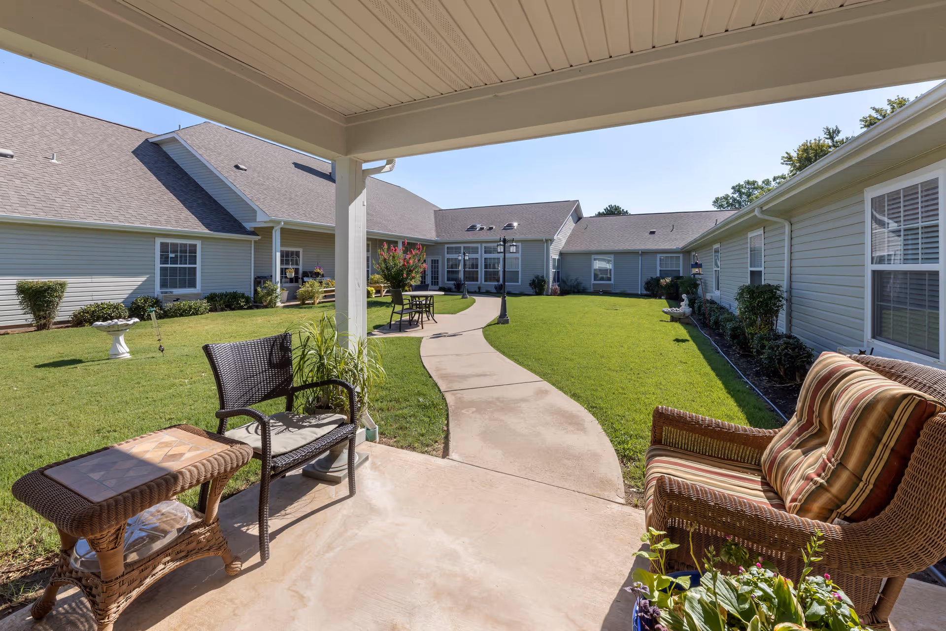 Covered patio area with wicker chairs and a small table overlooking a grassy courtyard with a winding concrete path, surrounded by single-story buildings with gray siding and white trim under a clear blue sky.