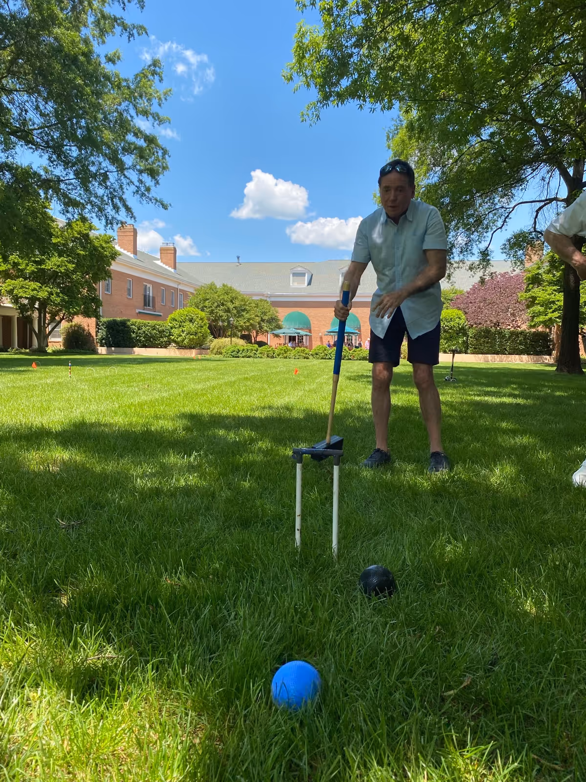 A man playing croquet on a grassy lawn with a brick building and trees in the background under a blue sky with some clouds.
