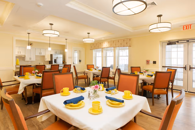 Bright communal dining room with tables set with yellow dishes, blue napkins, and orange chairs.