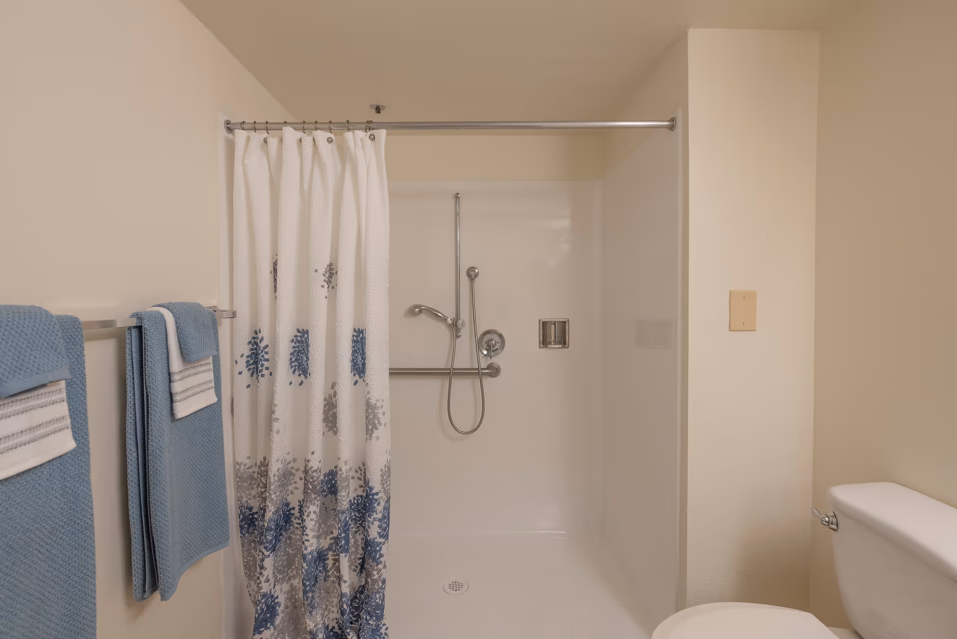 Bathroom with a white shower area featuring a silver showerhead and grab bar, a white shower curtain with blue and gray patterns, blue towels hanging on a towel rack, and a white toilet partially visible on the right.