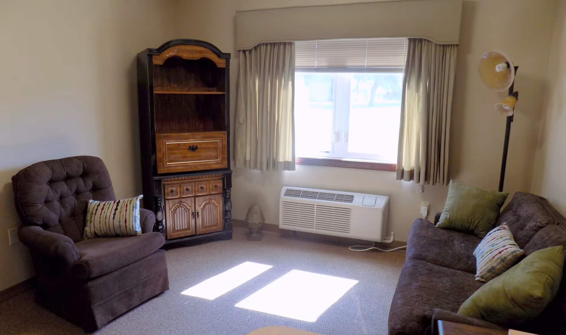 A cozy living room with a brown cushioned armchair and a matching sofa, both adorned with decorative pillows. There is a wooden cabinet with shelves and drawers against the wall, a window with beige curtains letting in natural light, and a floor lamp with two adjustable lights in the corner. The room has beige walls and carpeted flooring.