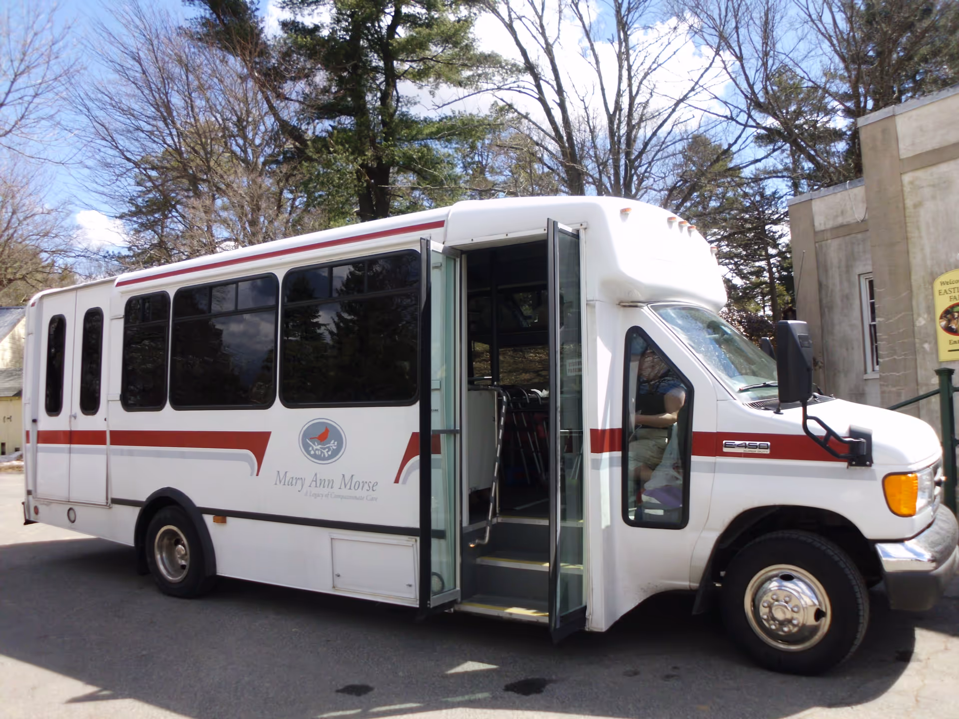 White shuttle bus with red stripes and the logo and name 'Mary Ann Morse A Legacy of Compassionate Care' on the side, parked outside near trees and a building. The bus door is open, showing steps inside.