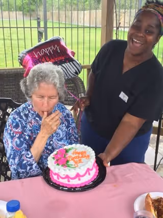 An elderly woman sitting at a table with a pink tablecloth, about to blow out the candle on a birthday cake decorated with pink and white icing and flowers. A smiling caregiver stands next to her holding the cake. In the background, there is a 'happy birthday' balloon and a fenced outdoor area with green grass.