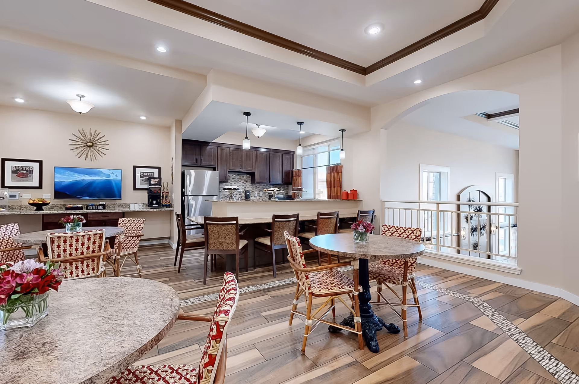 A bright and spacious dining area in an assisted living facility featuring round tables with floral centerpieces and patterned chairs. The room has wood-look tile flooring with a decorative inlay, a kitchen area with dark wood cabinets, stainless steel refrigerator, and a counter with bar stools. The walls are light-colored with crown molding, and there are pendant lights hanging over the kitchen counter. A TV and coffee bar are visible on the far wall.