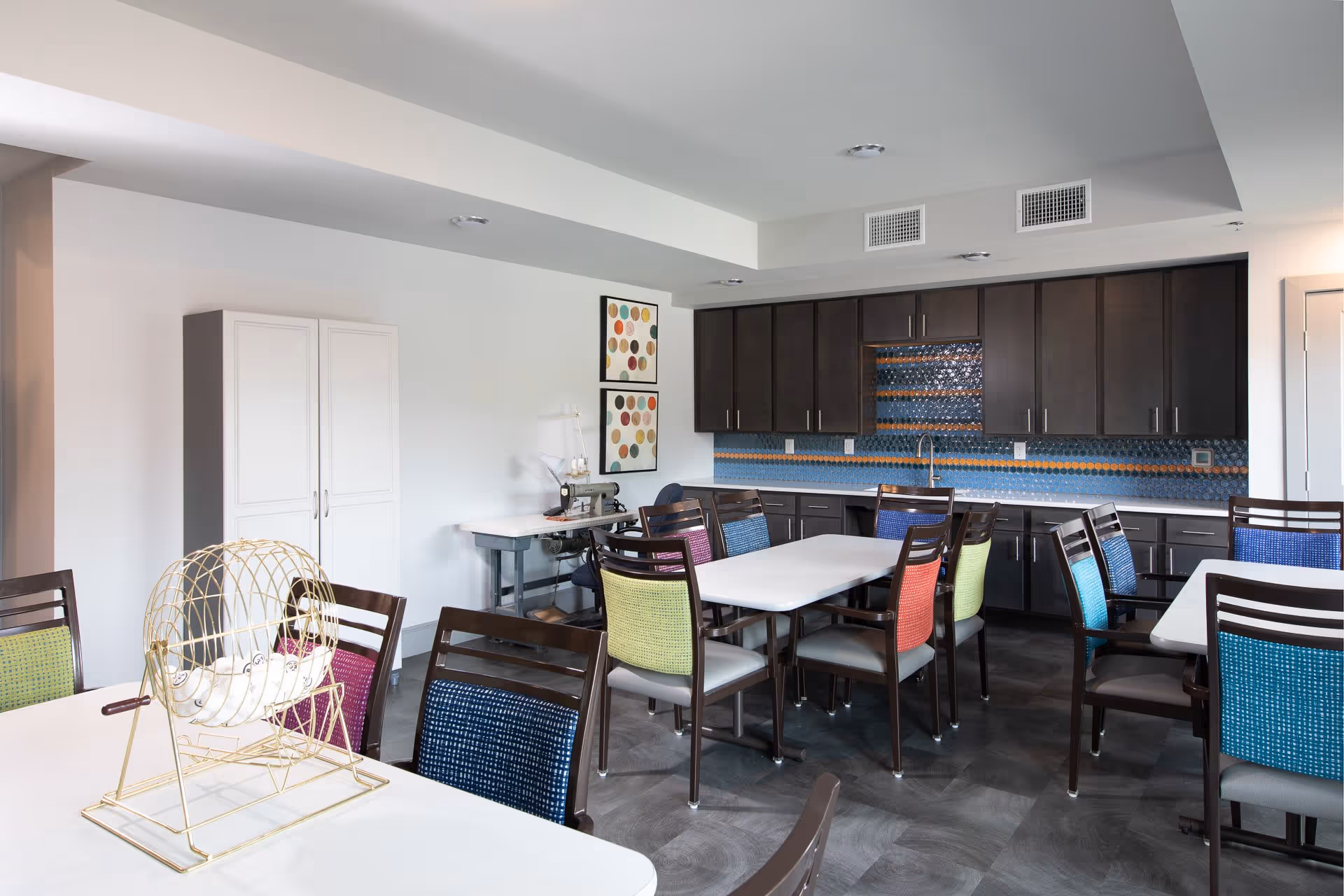 A bright and modern activity room with white tables and colorful chairs arranged around them. There is a gold bingo cage on one table. The back wall features dark cabinets with a blue and orange tiled backsplash and a sink. Two framed abstract artworks hang on the white wall next to a sewing machine on a table.
