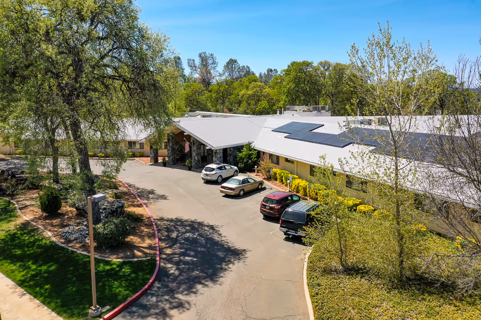 Front entrance of a single-story nursing center with a covered drop-off, parked cars, solar panels on the roof, and surrounding trees and landscaping.