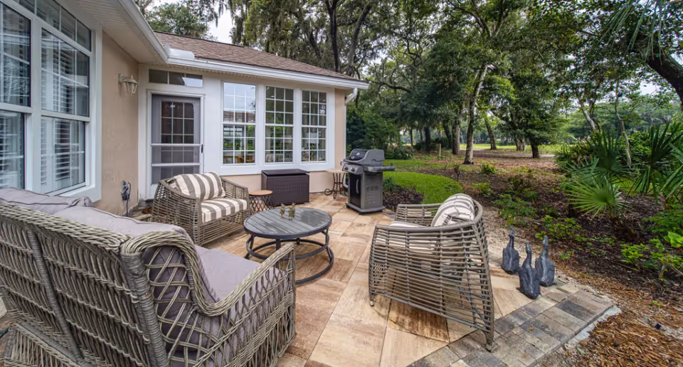 Patio seating area with wicker chairs and sofa, round coffee table, barbecue grill, and surrounding trees and landscaping.