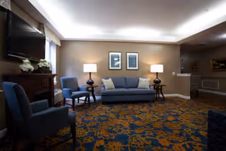 A cozy living room area with a blue sofa centered against a beige wall, flanked by two side tables each with a lamp. Two framed pictures hang above the sofa. There are two blue armchairs positioned on either side of the room. A large flat-screen TV is mounted on the wall to the left above a wooden cabinet with decorative flowers. The floor is covered with a patterned carpet in blue and gold tones. The room is softly lit with ceiling lights and lamps.