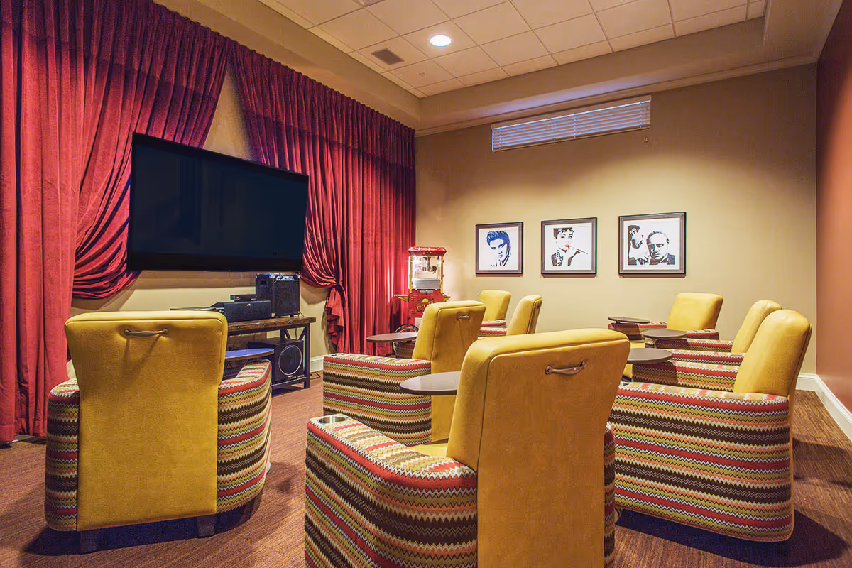 Small screening room with yellow and patterned armchairs facing a large wall-mounted TV, red curtains, a popcorn machine, and framed portraits on the wall.