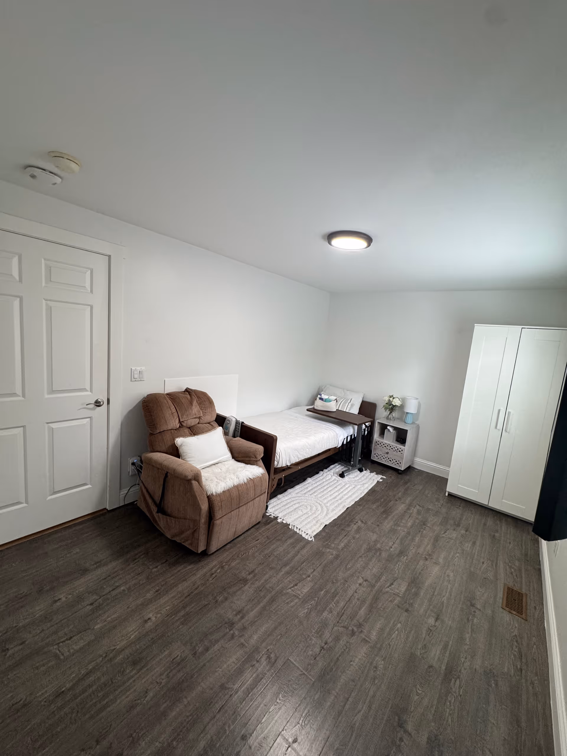 A simple, clean bedroom with a single bed against the wall, a brown recliner chair with a white pillow and throw, a small white nightstand with a lamp and flowers, a white wardrobe, and wood flooring. The walls and ceiling are white, and there is a closed white door on the left side.