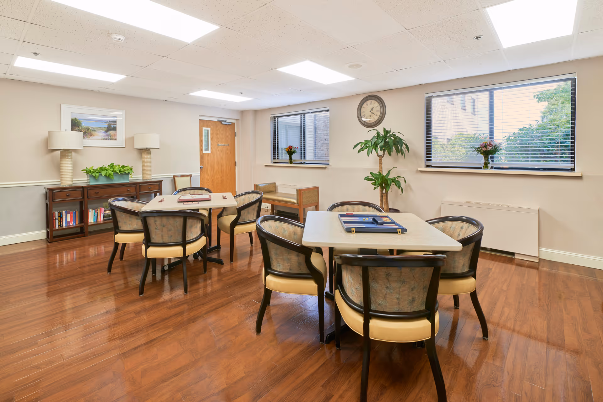 A well-lit common room with two tables surrounded by chairs, a wooden sideboard with two lamps and a plant, a wall clock, two windows with blinds, and a potted plant in the corner. The room has wooden flooring and neutral-colored walls.