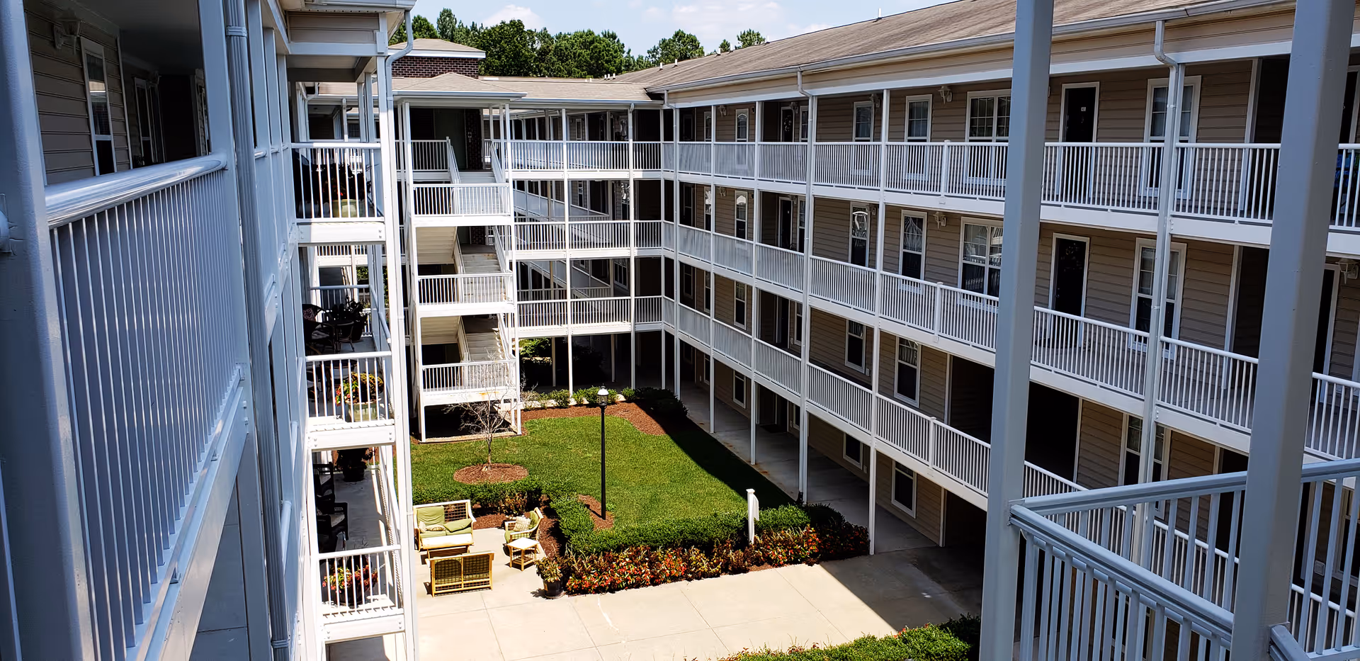 Multi-story apartment courtyard with white balcony railings, walkways around a grass lawn and a small outdoor seating area.