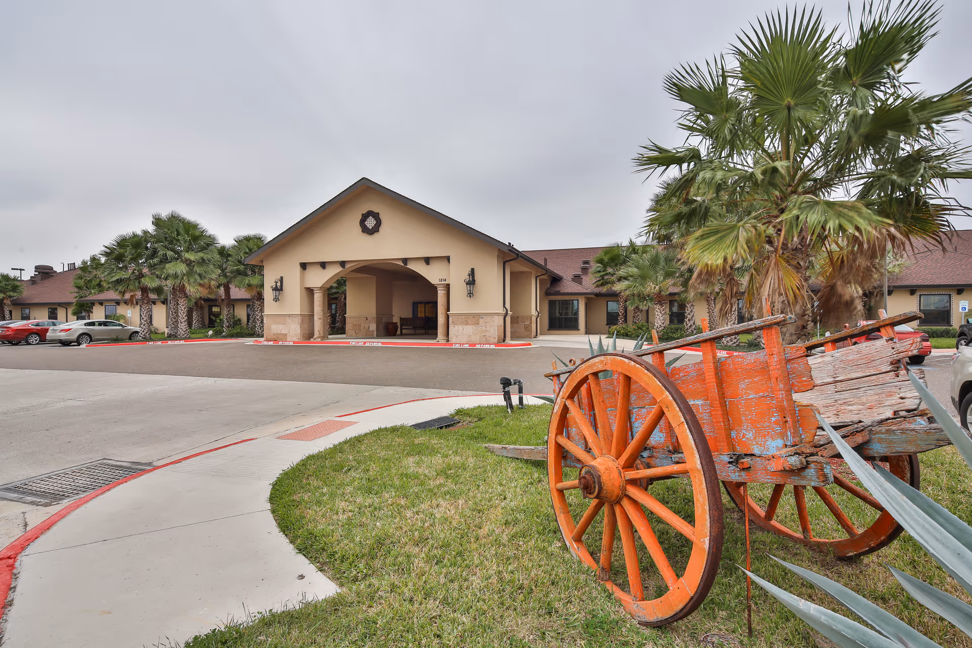 Exterior view of a senior living facility entrance with a covered porte-cochère, palm trees, parked cars, and a weathered orange wooden cart on the front lawn.