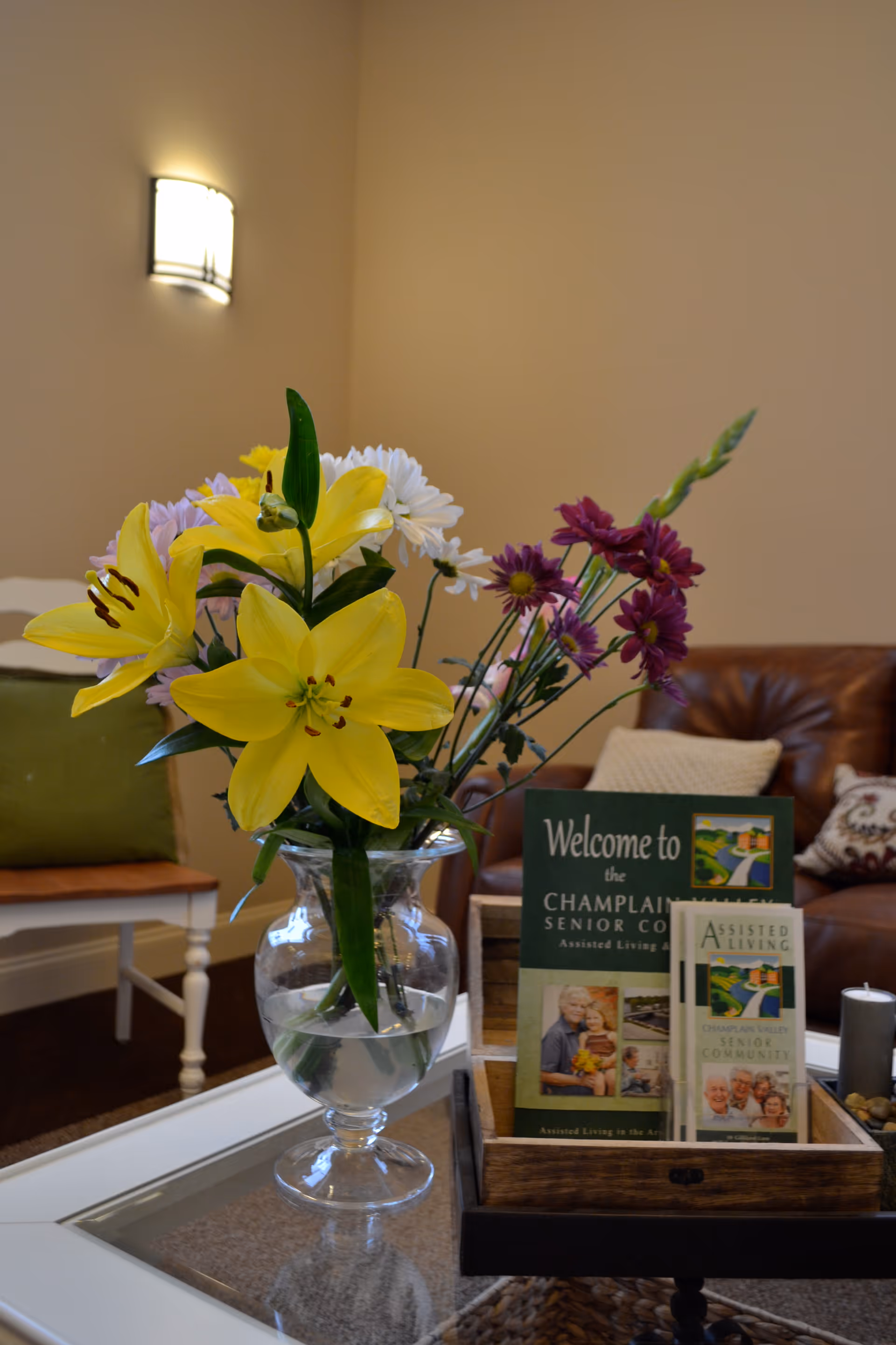 A vase with yellow lilies and other colorful flowers on a glass coffee table in a cozy living room. Behind the vase, there is a wooden tray holding brochures for Champlain Valley Senior Community. The background includes a brown leather couch with pillows and a white chair with a green cushion against a beige wall with a wall-mounted light fixture.