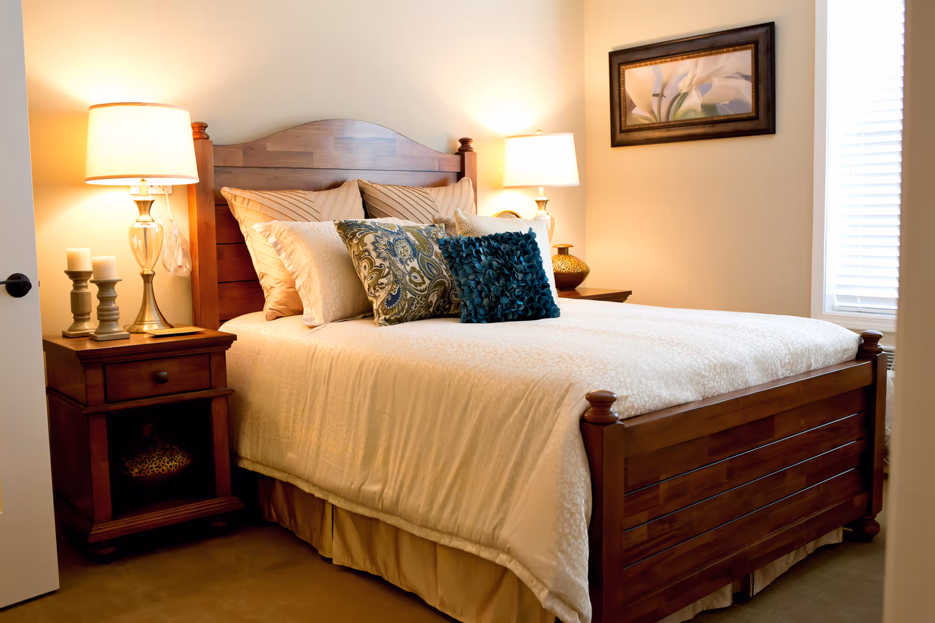 A cozy bedroom with a wooden bed frame and headboard, neatly made with cream-colored bedding and decorative pillows in blue and patterned designs. There are two wooden nightstands on either side of the bed, each with a lamp and decorative items. A framed floral artwork hangs on the wall above the bed, and a window with blinds is visible on the right side.