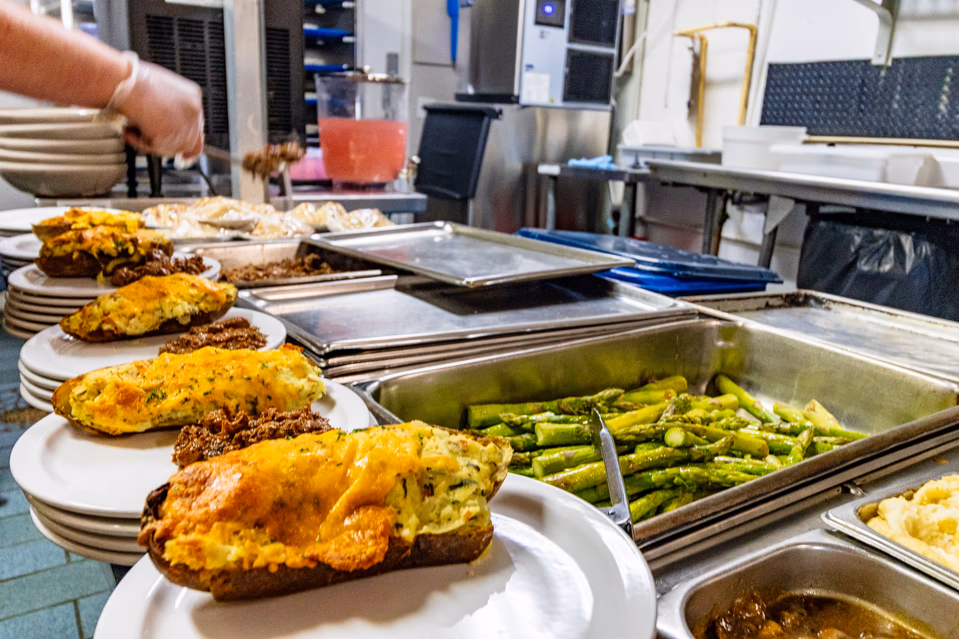 A kitchen serving line with plates of food including twice-baked potatoes topped with melted cheese and a side of ground meat, alongside trays of cooked asparagus and other hot dishes. A person is seen serving food in the background.