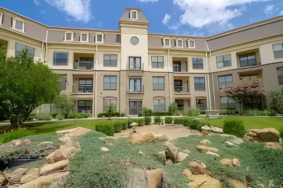 Front exterior of a multi-story senior living building with a landscaped courtyard and rock garden under a blue sky.