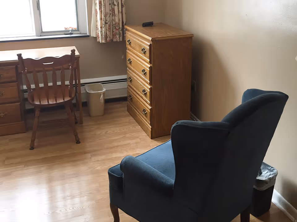 Small furnished room with an upholstered armchair, wooden dresser and desk by a window.