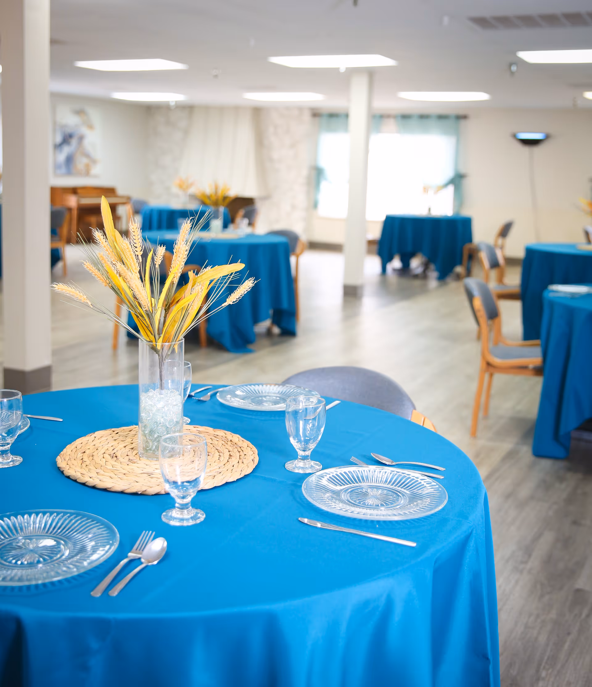 Dining room with round tables covered in blue tablecloths set with glass plates, utensils, and a vase centerpiece.
