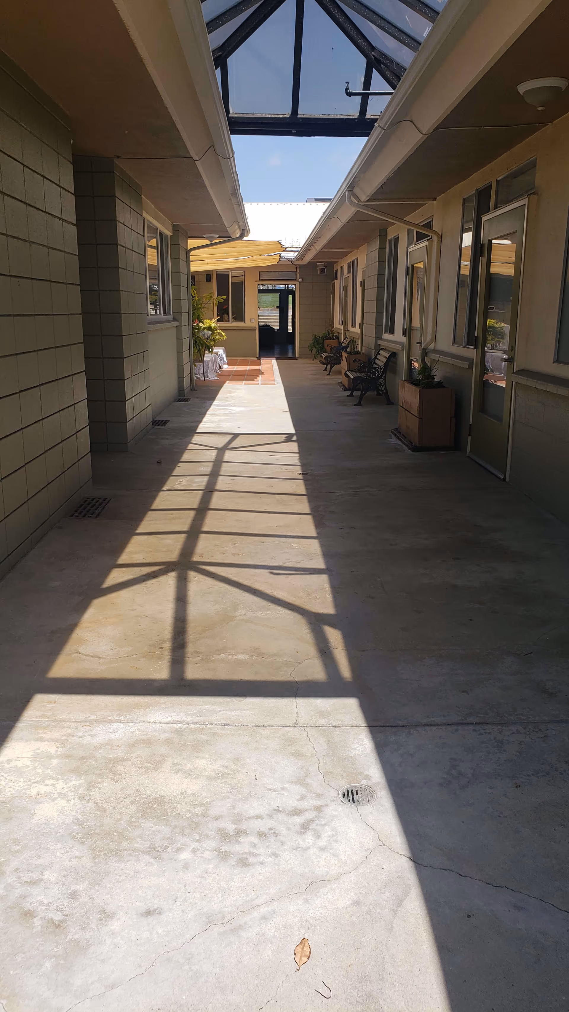 A covered outdoor walkway with a glass roof casting shadows on the concrete floor. The walkway is flanked by building walls with windows and doors on the right side, and a bench and potted plants along the path. At the far end, there is an open doorway leading to another area with more sunlight.
