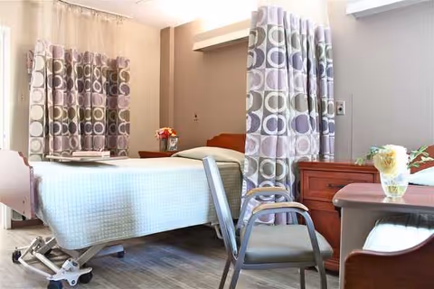 A senior living facility bedroom with a hospital-style bed, privacy curtains, dresser, and chair.