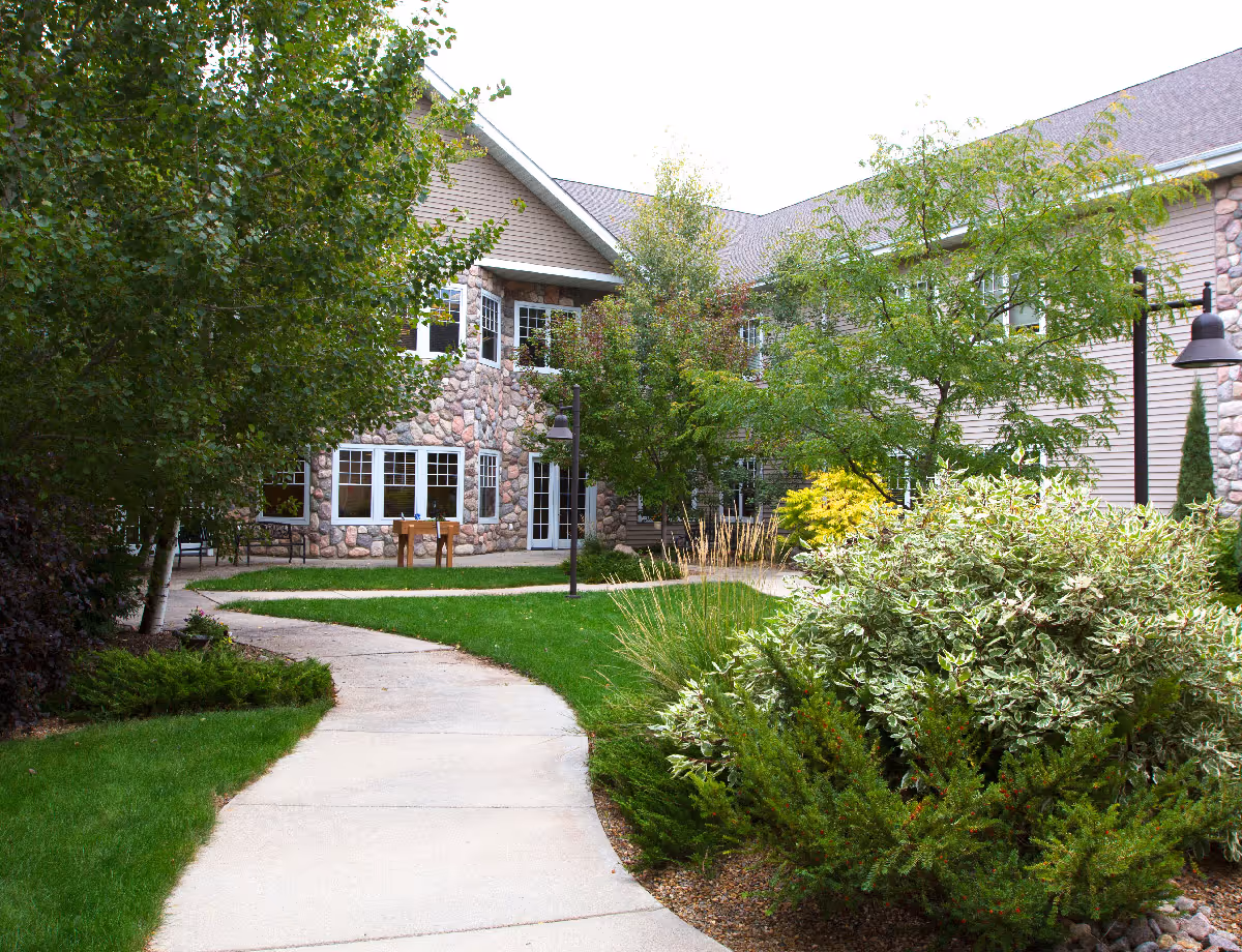 Curved sidewalk through a landscaped courtyard leading to the stone-faced exterior of a two-story senior living building.