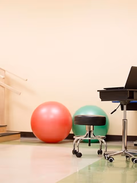 A room with a red and a green exercise ball near a wall, a black rolling stool, and part of a black desk with a laptop on it. There are wooden handrails on the left side and a green and beige tiled floor.